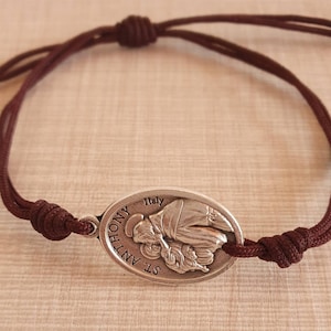 Puede incluir: Un brazalete de cordón marrón con un dije ovalado de plata que representa a San Antonio de Padua sosteniendo al niño Jesús. El dije está inscrito con "St. Anthony" e "Italia".