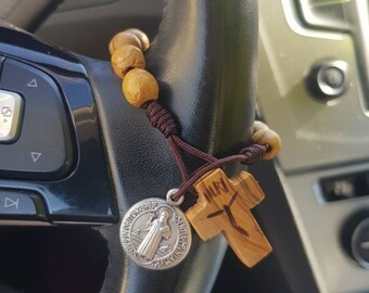 Könnte beinhalten: Ein braunes Holzrosenkranz mit einem silbernen Anhänger und einem Holzkreuz, das am Lenkrad eines Autos hängt. Der Anhänger zeigt eine Heilige.
