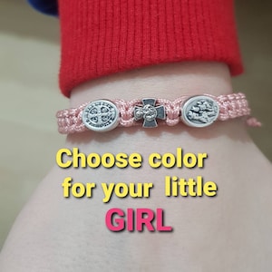 May include: A pink braided bracelet with silver charms, including a cross and a medal. The text "Choose color for your little GIRL" is written below the bracelet.