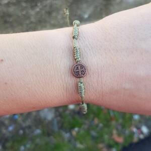 May include: A green and brown braided bracelet with a round metal charm featuring a cross design.