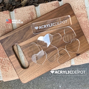 May include: A wooden cutting board with a handle and a clear acrylic heart-shaped stencil. The stencil is labeled "ACRYLICDEPOT" and "Made in America".