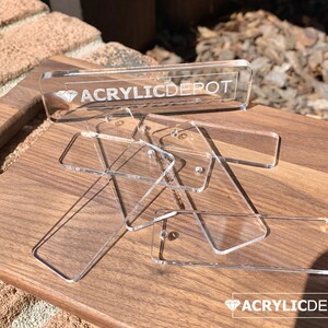 May include: A close-up of several clear acrylic rectangular shapes with rounded corners. The shapes are stacked on top of each other on a wooden surface. The top shape has the text "ACRYLICDEPOT" and a diamond symbol engraved on it.