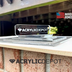 May include: A clear acrylic display stand with multiple layers. The stand features the text "ACRYLICDEPOT" in black lettering on a black diamond shape. The stand is made in the USA, as indicated by the red, white, and blue American flag with the text "MADE IN AMERICA".