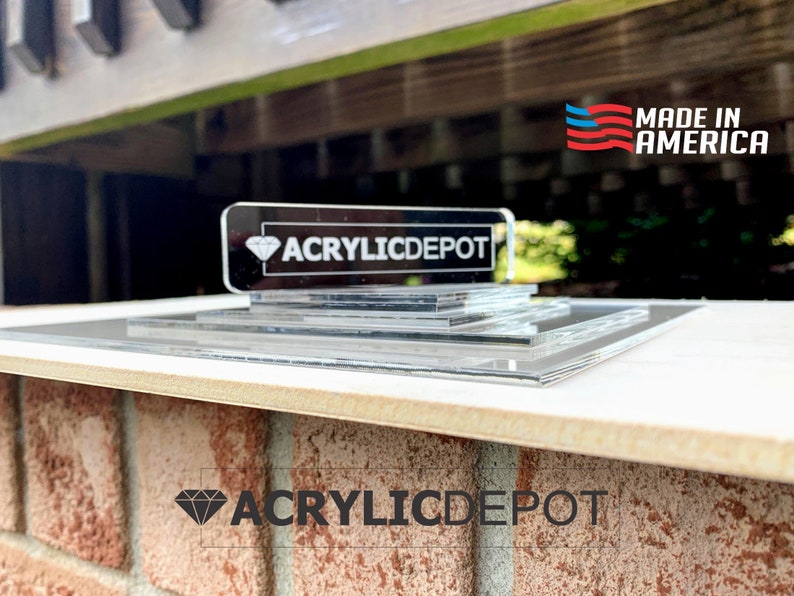 May include: A clear acrylic display stand with multiple layers. The stand features the text "ACRYLICDEPOT" in black lettering on a black diamond shape. The stand is made in the USA, as indicated by the red, white, and blue American flag with the text "MADE IN AMERICA".