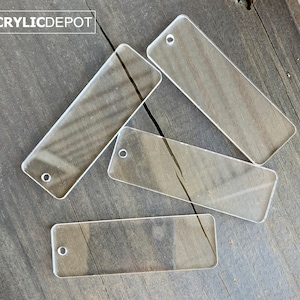 May include: Four clear acrylic rectangular tags with a hole punched in the top. The tags are on a wooden surface.