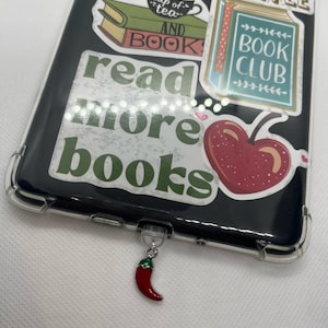May include: A black phone case with various colourful stickers featuring books, cherries, and the text "read more books". A red chilli pepper charm hangs from the bottom of the case.