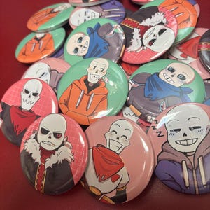 Puede incluir: Una colección de botones redondos y coloridos con varios diseños de personajes de dibujos animados. Los botones muestran personajes con rasgos esqueléticos, que llevan sudaderas con capucha, bufandas y otros accesorios. Los botones son en tonos de rojo, verde, naranja y azul.
