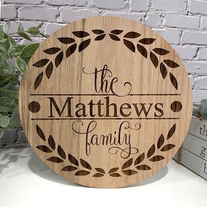 May include: A round wooden sign with the words "The Matthews Family" engraved in a decorative font. The sign is surrounded by a laurel wreath design and has a natural wood grain finish.