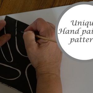 May include: A hand paints a black and white abstract pattern on a ceramic tile. The tile has a unique hand painted pattern.