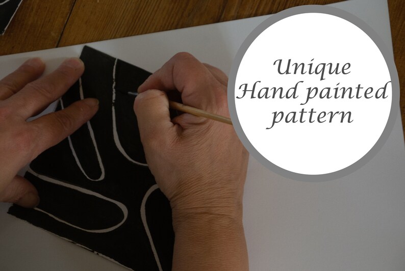May include: A hand paints a black and white abstract pattern on a ceramic tile. The tile has a unique hand painted pattern.