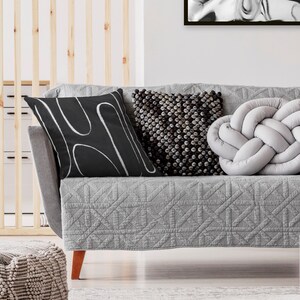 May include: A gray sofa with a quilted cover and three throw pillows. The pillows are black with white abstract lines, gray with a textured surface, and a light gray knotted pillow.