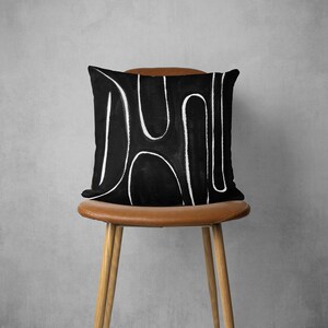 May include: A black and white throw pillow with an abstract design. The pillow is sitting on a brown chair.