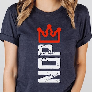 May include: Heather navy blue t-shirt with a red crown design above the word "NOP" in white, distressed font. The shirt is a crew neck with rolled-up sleeves. A circular logo in the bottom right corner reads "Heather Navy".