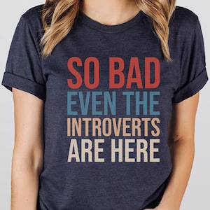 May include: Heather navy blue t-shirt with the text "SO BAD EVEN THE INTROVERTS ARE HERE" in red, blue, and beige. The shirt has rolled-up sleeves and is made of a soft material. The shirt is a unisex style.