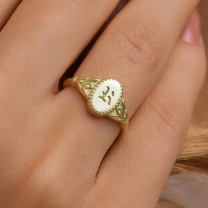 Puede incluir: Un anillo de sello de oro con una parte superior ovalada que presenta un diseño grabado. El anillo se lleva en un dedo.