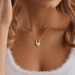 Peut inclure: Un collier doré avec un pendentif en forme de cœur. Le pendentif porte une petite lettre. Le collier est porté par une personne vêtue d'un haut blanc. L'arrière-plan est de couleur beige uni.