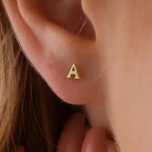 Peut inclure: Une boucle d'oreille en or avec la lettre 'A' en forme de clou. La boucle d'oreille est portée au lobe gauche d'une personne aux cheveux blonds.