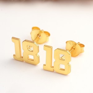 May include: Gold-toned earrings with the number "18" on each earring. The earrings are stud style.