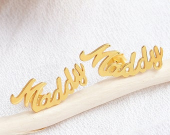 May include: A pair of gold-colored earrings with the word "Maddy" written in a cursive font.