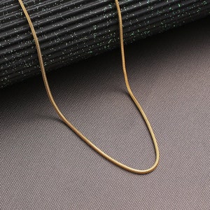 May include: A gold snake chain necklace on a black textured surface.