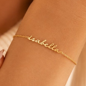 May include: A gold-toned chain bracelet featuring the name isabella written in a delicate, cursive script font. The bracelet is composed of a thin, polished metal chain that connects to the central nameplate, creating a continuous, elegant design.