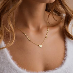 May include: A delicate gold necklace featuring the names "Emily" and "Amanda" in cursive script, connected by a small heart pendant. The necklace is worn around the neck, showcasing the personalized design.