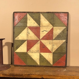 Puede incluir: Arte mural cuadrado de madera con un diseño geométrico de bloque de colcha. El bloque presenta triángulos en colores rojo, verde y crema. La madera tiene un aspecto envejecido y desgastado, con desgaste visible.