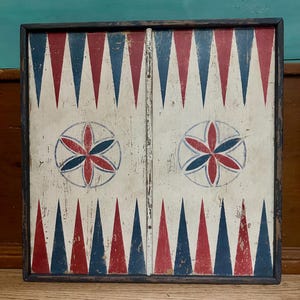 May include: A vintage backgammon board with a distressed, aged appearance. The board features a white background with alternating red and blue triangles. Two circular floral designs are centered on the board. The frame is dark brown.