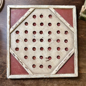 May include: Square, antique-style game board with a cream-colored frame and a red center. The center features a diamond-shaped design with numerous small holes. The frame has a distressed finish, giving it a vintage appearance. The board is likely for a peg game.