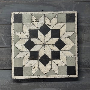 May include: Square, distressed wooden wall art featuring a geometric starburst design. The artwork incorporates black, white, and gray tones, with a weathered, vintage aesthetic. The piece is approximately 12 inches square.