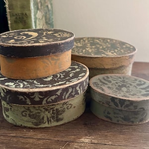 May include: A set of three oval-shaped boxes with lids, featuring distressed finishes in shades of brown, green, and dark gray. Each box displays a decorative, repeating pattern on the lid and sides, showing signs of wear and texture.
