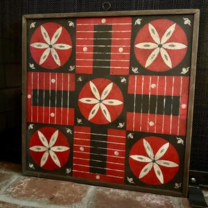 May include: A square wooden game board featuring a distressed finish with a black background and red rectangular sections. The design includes five red circles, each painted with a white six-petaled flower motif, and white lines throughout.