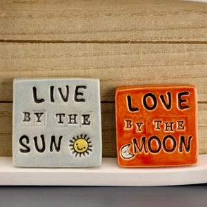 Könnte beinhalten: Zwei quadratische Keramikfliesen mit inspirierenden Zitaten. Eine Fliese ist hellblau mit den Worten "LIVE BY THE SUN" und einer Sonnenillustration. Die andere Fliese ist orange mit den Worten "LOVE BY THE MOON" und einer Mondillustration.