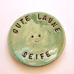 Könnte beinhalten: Eine runde, helltürkisfarbene Keramik-Seifenschale mit den Worten "GUTE LAUNE SEIFE" in schwarzer Schrift. Die Schale hat ein lächelndes Gesicht mit zwei Augenlöchern und einem roten, gebogenen Mund. Die Schale hat eine gesprenkelte Textur.