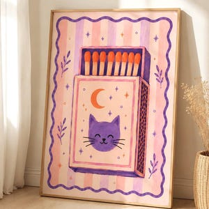 May include: A framed wall art print featuring a matchbox illustration on a pink and lavender striped background. The matchbox displays a purple cat face, an orange crescent moon, and stars, with orange-tipped matches protruding from the top.