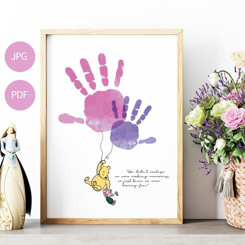 May include: A framed print featuring two handprints in watercolor shades of pink and purple. Winnie the Pooh and Piglet are depicted hanging from the handprints. The quote "We didn't realize we were making memories, we just knew we were having fun" is included.