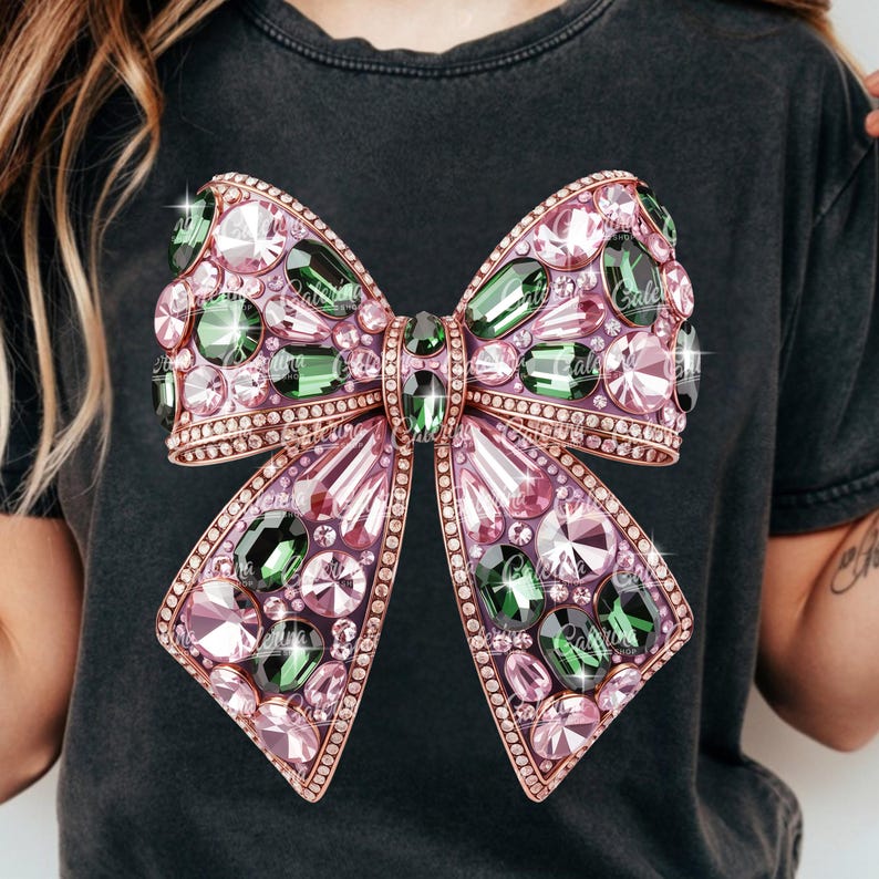 May include: A dark gray t-shirt featuring a large, ornate bow design. The bow is embellished with pink and green gemstones, outlined with a gold-colored trim. The design has a sparkling effect, adding to its glamorous appearance.