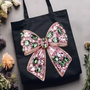 May include: Black tote bag featuring a large, ornate bow design. The bow is embellished with pink, green, and clear gemstones set in a gold-colored metal frame. The bag has black handles and is likely made of fabric.