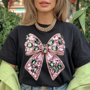 May include: Black t-shirt featuring a large, embellished bow design. The bow is adorned with pink and green gemstones, creating a sparkling effect. A gold chain necklace is visible at the neckline. The shirt is a casual wear item.