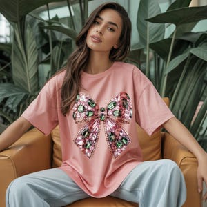 May include: A light pink t-shirt featuring a large, embellished bow design. The bow is adorned with green and pink gemstones, creating a sparkling effect. The shirt has short sleeves and a relaxed fit.