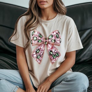 May include: A beige t-shirt featuring a large, ornate bow design. The bow is embellished with pink and green gemstones, creating a sparkling effect. The shirt is a casual style, suitable for everyday wear.