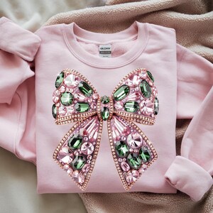 May include: A light pink sweatshirt featuring a large, jeweled bow design. The bow is adorned with pink, green, and clear crystals, outlined in a gold-colored metal. The sweatshirt has a crew neck and long sleeves.