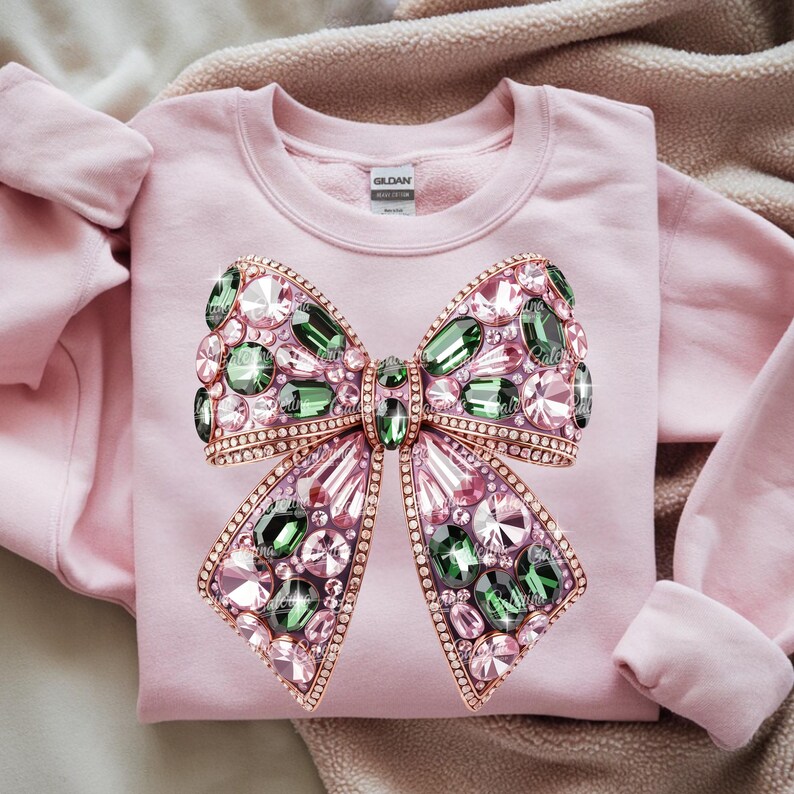 May include: A light pink sweatshirt featuring a large, jeweled bow design. The bow is adorned with pink, green, and clear crystals, outlined in a gold-colored metal. The sweatshirt has a crew neck and long sleeves.