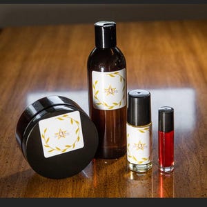 Puede incluir: Una colección de productos de belleza, que incluye un tarro, una botella y dos viales más pequeños, dispuestos sobre una superficie de madera. Cada envase presenta una etiqueta blanca con un diseño de hojas doradas y una estrella. Los viales contienen un líquido rojo.