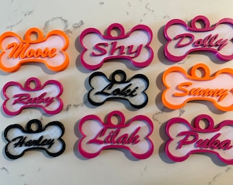 May include: Nine personalized dog bone-shaped tags with different names. The tags are made of white plastic with a colored border and the names are printed in a bold, cursive font. The colors of the borders are pink, orange, and black.
