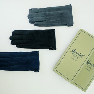 Puede incluir: Tres pares de guantes de gamuza en diferentes colores: azul oscuro, negro y gris. Los guantes están dispuestos sobre una superficie blanca con dos cajas etiquetadas como "Macahrel London UK Brand" en el fondo.
