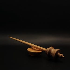 May include: A light brown wooden hair stick with a smooth, tapered shaft and a decorative, turned finial featuring rounded ridges and a pointed tip, resting horizontally on a small, matching circular wooden stand against a black background.