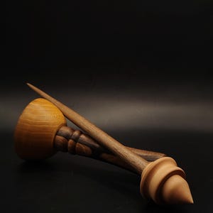 Op de afbeelding: Een houten spintol met een lichtgekleurde spintolwervel in een gladde, ronde komvorm, verbonden aan een donkere, gedraaide houten schacht die uitloopt in een scherpe punt, vergezeld door een apart lichtgekleurd houten wervelonderdeel.