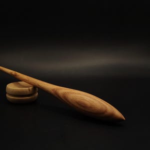 May include: A light brown wooden hair stick with a smooth, tapered, and elongated spindle shape. The wood grain is visible, showing natural patterns and swirls throughout the length of the stick. It rests against a small, round wooden base.