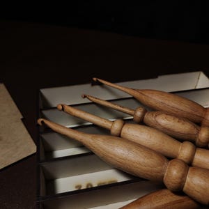 Może przedstawiać: Kolekcja drewnianych szydełek, każde z gładkim, zaokrąglonym uchwytem i zakrzywionym końcem haka. Szydełka ułożone są w przegródkowej skrzynce, ukazując naturalne usłojenie drewna i ciepłe odcienie. Obraz jest dobrze oświetlony, podkreślając kunszt wykonania.